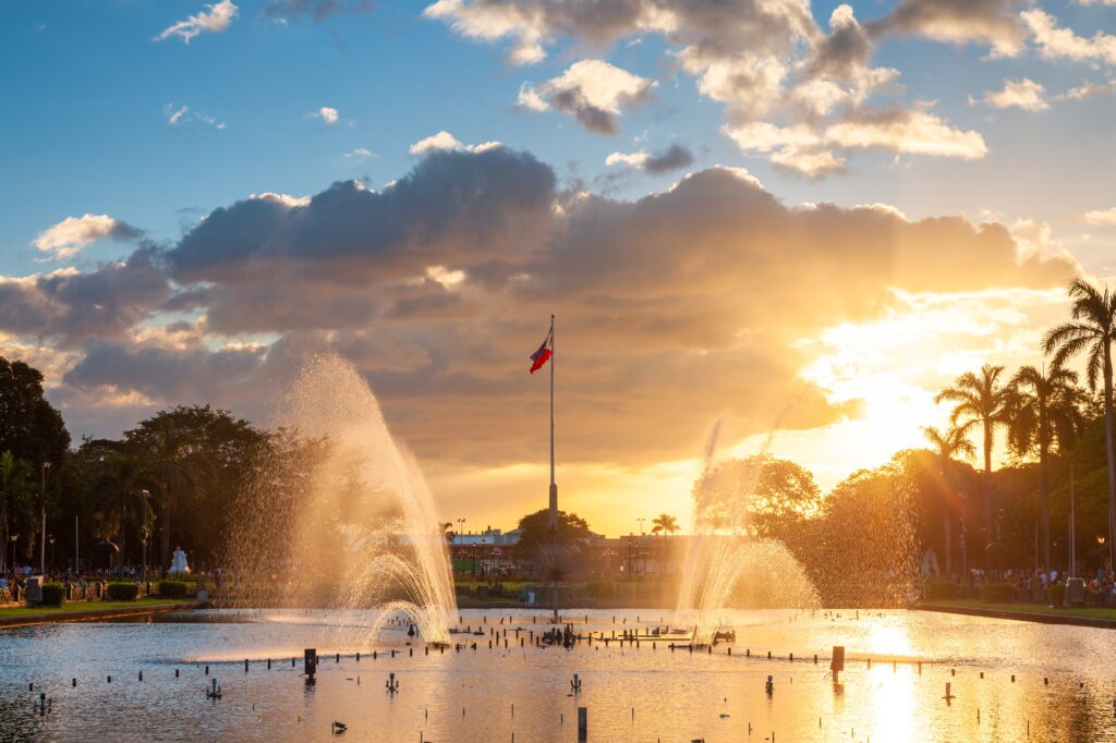 View of a fountain in Rizal Park with a flag of the Philippines under beautiful sunset light